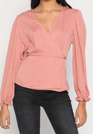 Mujer que lleva una blusa cruzada de manga larga color rosa polvo con mangas abullonadas y pantalones negros ajustados, mostrada desde el cuello hasta las caderas.