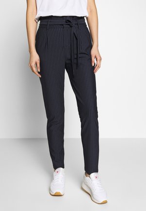 Vrouw in marineblauwe streepjes broek met paperbag taille en gestrikt riempje, gecombineerd met witte sneakers, tegen een effen achtergrond.