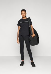 Zwarte katoenen t-shirt met "Champion" logo, zwarte slim-fit broek en zwarte sneakers. Draagt een zwarte sporttas met een gladde textuur.