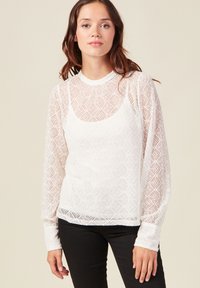 Weiße Spitzenbluse mit langen Ärmeln aus diamantgemustertem Stoff, Rundhalsausschnitt und transparenter Textur, über einem eng anliegenden Top getragen.