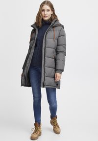 Gray puffer coat with a hood, featuring orange drawstrings, two side pockets, and a zip front. Worn over a navy sweater and blue jeans.