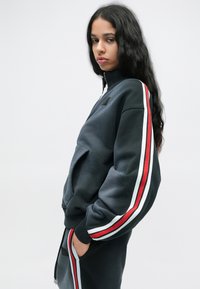 Γυναίκα με μακριά σκούρα μαλλιά ντυμένη με μαύρη φόρμα με κόκκινες και λευκές ρίγες στα μανίκια και το παντελόνι, στέκεται με το ένα χέρι στην τσέπη.