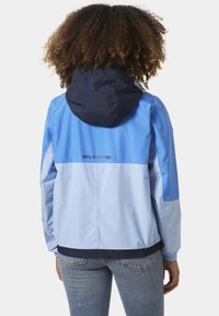 Lichtblauwe windbreaker jas met een marineblauwe capuchon, elastische manchetten en een trekkoord onderaan. Het heeft een zwart logo op de bovenrug.