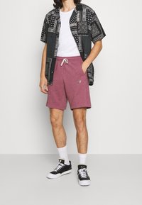 Osoba v černé vzorované košili s krátkým rukávem přes bílém tričku, v bordových Nike šortkách, bílých ponožkách a černých Nike teniskách.