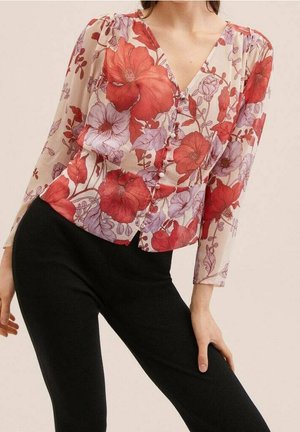 Kobieta w przejrzystej, kwiecistej bluzce z czerwonymi i lawendowymi kwiatami oraz czarnych dopasowanych spodniach, pozująca z jedną ręką na udzie.