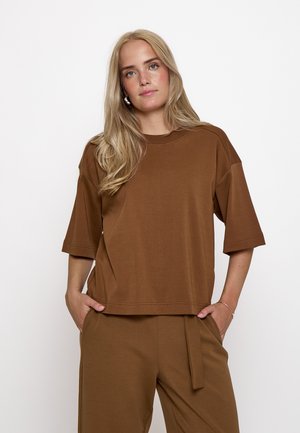 Blonde Frau mit offenem Haar, trägt ein braunes übergroßes Kurzarmshirt und passende Hosen, Hände in den Taschen.