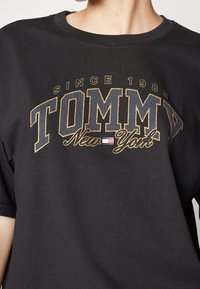 Černé bavlněné tričko s logem ve zlaté a modré barvě s prasklým tiskem, které říká "TOMMY New York" a nápisem "Since 1985" nahoře, kulatý výstřih, krátké rukávy.
