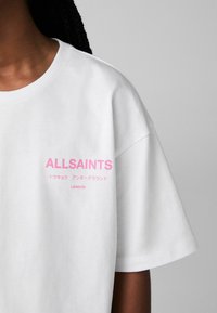 Balta medvilnė marškinėliai su trumpomis rankovėmis, su rožine užrašu "ALLSAINTS" ir papildomu tekstu japonų kalba bei "LONDON" šalia apatinio krašto.