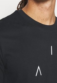 Černé bavlněné tričko s kulatým výstřihem. Obsahuje bílé grafické texty a hladký povrch. Detailní záběr oblasti ramene.