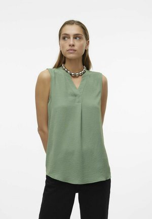 Haut sans manches vert clair avec un col en V, finition texturée et ourlet arrondi. Porté avec un grand collier de perles métalliques.