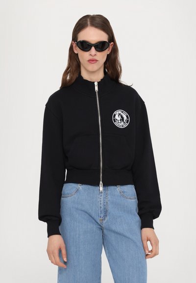 Black cropped sweatshirt with a high collar, zip front, and front pocket, featuring a circular white logo on the left chest. Paired with blue jeans.