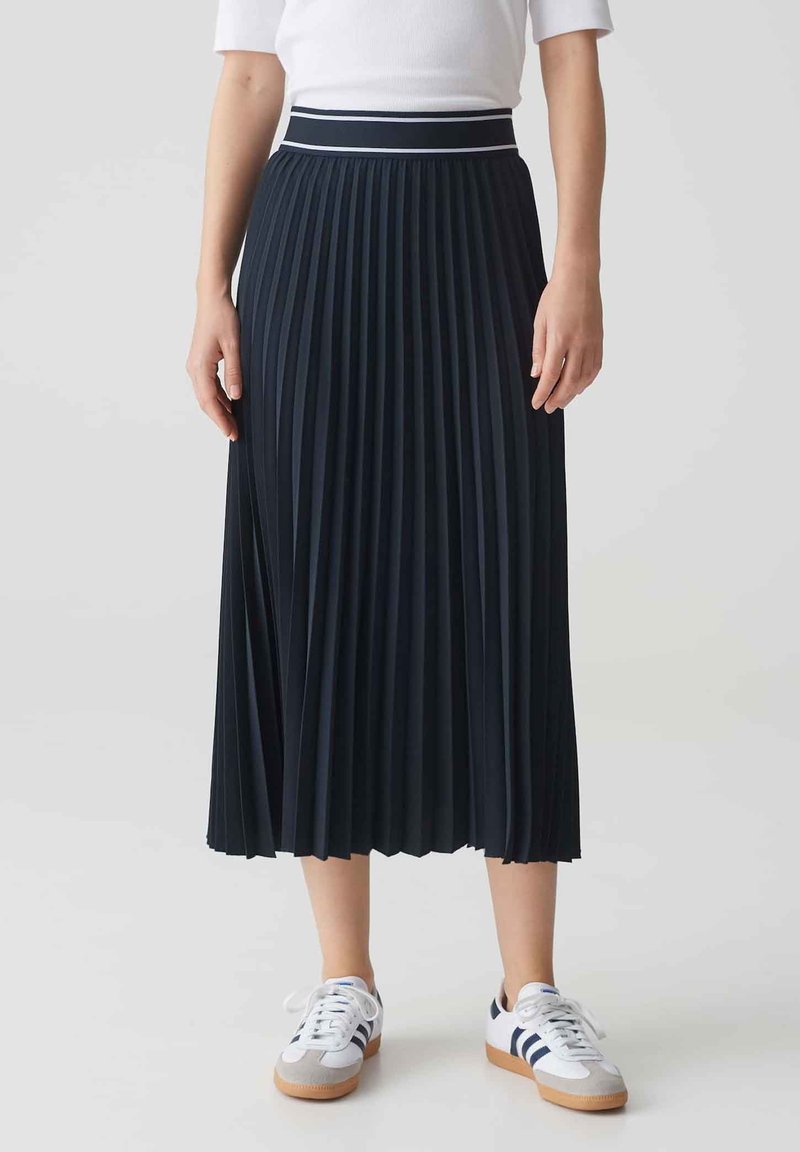 Jupe midi plissée bleu marine en tissu texturé, dotée d'une taille blanche contrastante. Assortie à des baskets blanches et des accents bleus.