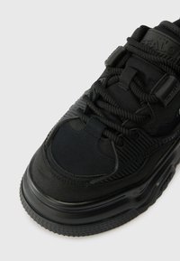 Pantofele de sport negre, cu talpă groasă și texturată, limbă căptușită și șireturi rotunde, prezentate dintr-un unghi superior pe fundal alb.