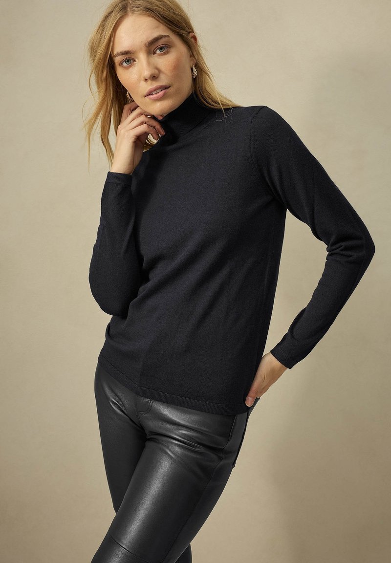 Schwarzer Rollkragenpullover aus weichem Strickstoff mit langen Ärmeln und taillierter Passform. Kombiniert mit schwarzen Lederhosen.