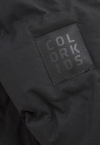 Svart tyg med fin struktur, med ett rektangulärt, svart gummimärke präglat med "COLOR KIDS" i fetstil.