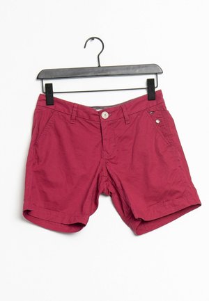 Shorts en coton rouge avec fermeture à bouton, poches latérales et ourlets retroussés, suspendus à un cintre en plastique noir sur fond blanc.
