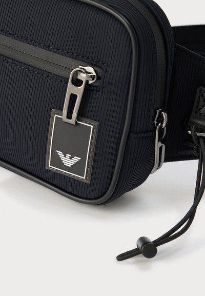 Κοντινό πλάνο ενός μαύρου τσάντας ώμου με υφή, ασημένια φερμουάρ, ρυθμιζόμενο λουρί και ετικέτα με λογότυπο που απεικονίζει έναν στυλιζαρισμένο αε τό.