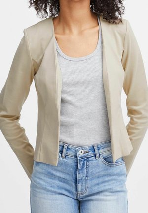 Woman wearing a beige open blazer, light gray ribbed tank top, and high-waisted light blue jeans with hands behind back.