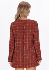 Abito in tweed rosso con un motivo a griglia, caratterizzato da maniche lunghe, un taglio dritto e una chiusura con bottone sulla schiena. Tessuto testurizzato con accenti colorati.