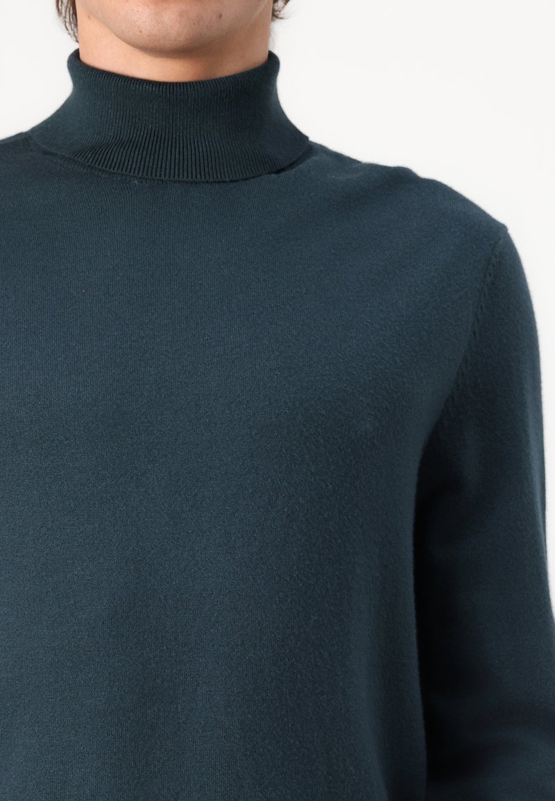 Pull en turtleneck bleu foncé, fabriqué dans un tissu doux, avec un col à côtes haut et une silhouette ajustée sans motifs ni ornements visibles.