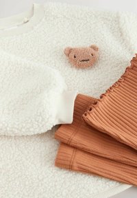 Biała, teksturowana bluza polarowa z naszywką przedstawiającą brązowego niedźwiedzia oraz prążkowanymi mankietami, w komplecie z trzema spodniami w kolorze rdzy o prążkowanej fakturze z falbanami.