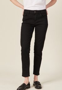 Czarne dżinsowe spodnie o slim fit, z klasycznym pięciopunktowym fasonem, zapięciem na guzik z przodu i długością do kostek. Noszone z czarnymi mokasynami.