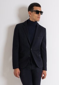Blazer blu navy con revers, due tasche frontali e chiusura a un bottone; indossato sopra un dolcevita blu a coste. Modello con occhiali da sole.