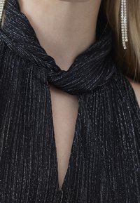 Top de tela negra con pliegues y brillo metálico, con un diseño de cuello halter y detalle en forma de llave en el pecho. Textura larga y suave.