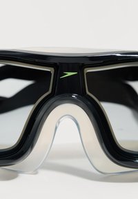 Μαύρα και διαυγή γυαλιά κολύμβησης με μεγάλα φακούς και ένα μικρό πράσινο λογότυπο στο κέντρο πάνω από τη γέφυρα της μύτης.