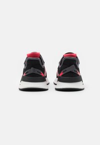 Par de zapatos deportivos en negro y rosa vistos desde atrás con suelas blancas y lengüetas rojas en el talón sobre un fondo blanco.