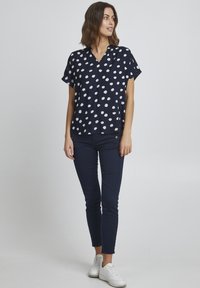 Granatowa bluzka z krótkim rękawem w białe kropki, dekolt w serek, luźny krój, zestawiona z dopasowanymi granatowymi spodniami i białymi trampkami.
