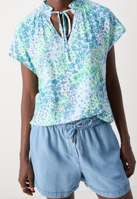 Blouse à manches courtes imprimé floral bleu et vert, avec un col froncé et un détail à nouer, associée à un short en denim clair.