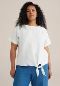 Top blanco de manga corta con un detalle de nudo atado en el dobladillo y un escote redondo. Llevado con pantalones azules de corte holgado. Tejido suave y ligero.
