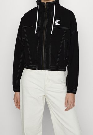 Czarna kurtka na suwak z białymi sznurkami i logo na piersi, noszona z białymi spodniami z wysokim stanem, kontrastujące białe szwy na kurtce.