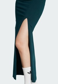 Zelená dlhá sukňa s vysokým bočným rozparkom, vyrobená z hladkého materiálu. Bielé ponožky s logom viditeľným na spodnej strane.