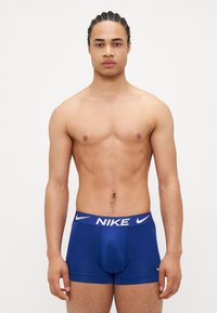 Boxeri Nike albaștri cu logo pe talie albă, fabrică netedă, design ajustat și croială de înălțime medie, prezentate pe un model masculin fără cămașă.