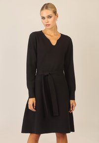 Schwarzes langärmliges Kleid mit V-Ausschnitt und Taillengürtel. Hergestellt aus weichem Material mit einem ausgestellt geschnittenen Rock. Sanfte Textur, minimalistisches Design.