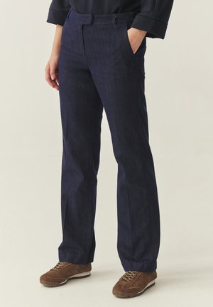 Person iført mørkeblå bukser med lige ben og brune ruskindssneakers, stående med den ene hånd i lommen mod en ensfarvet baggrund.