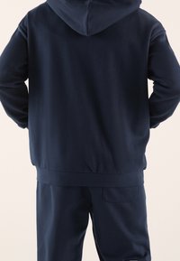 Felpa con cappuccio blu navy e pantaloni abbinati in morbido tessuto. Caratterizzata da cappuccio, polsini a costine e una tasca posteriore sui pantaloni. Design semplice.