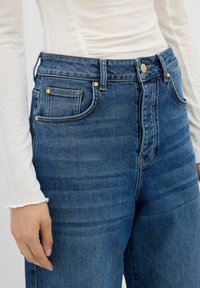 Jeans de mezclilla azul de tiro alto con corte recto, que cuentan con bolsillos frontales, acentos metálicos dorados y un acabado texturizado.