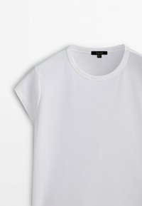 Maglietta a maniche corte bianca con scollo rotondo, poggiata piatta, che mostra la parte anteriore superiore con una piccola etichetta nera del marchio all'interno del colletto.