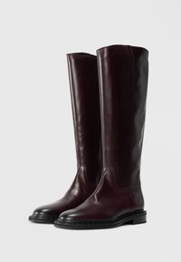 De grandes bottes en cuir marron foncé avec des bouts ronds et des semelles noires basses, se tenant côte à côte sur un fond clair et uni.