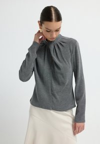 Grauer Strickoberteil mit hochgeschlossenem Kragen und Faltendetail, mit langen Ärmeln und lockerer Passform. Getragen über einem cremefarbenen Rock.