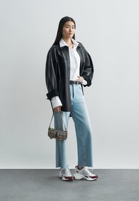 Čierna kožená oversize košeľa, biela zapínacia košeľa pod ňou, svetlomodré rifle s vysokým pásom, malá sivá kabelka a biele tenisky s červenými prvkami.