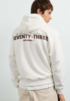 Jonge man draagt een witte hoodie met de tekst "Seventy-Three" en "Pepe Jeans" op de achterkant, kijkt opzij tegen een effen achtergrond.