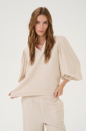 Haut beige texturé avec un décolleté en V et des manches bouffantes. Le tissu présente des motifs subtils, associé à un pantalon ample assorti.