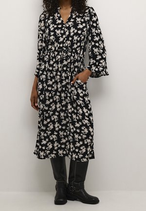 Femme portant une robe midi noire à imprimé floral blanc et manches longues, associée à des bottes noires montantes jusqu'aux genoux, debout devant un fond uni.