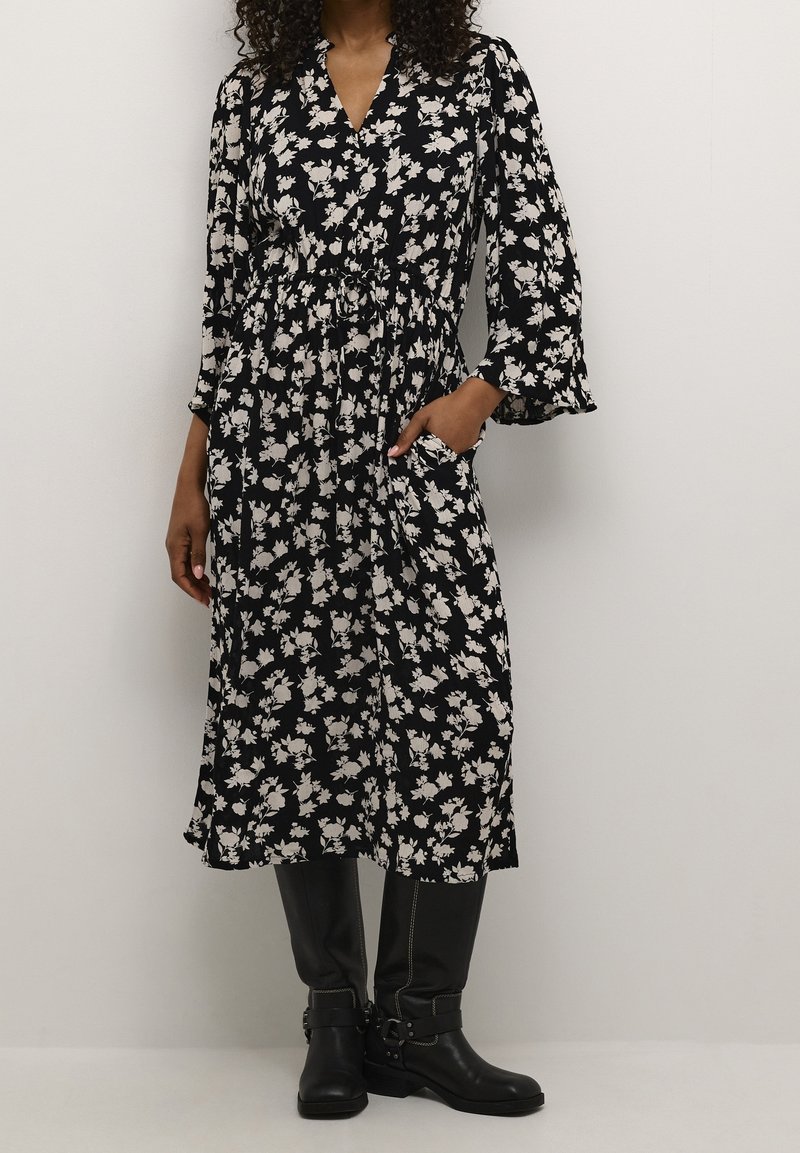 Femme portant une robe midi noire à imprimé floral blanc et manches longues, associée à des bottes noires montantes jusqu'aux genoux, debout devant un fond uni.