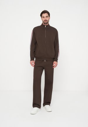 Homme debout portant un survêtement marron avec des bandes rouges et blanches aux manches et des baskets blanches sur un fond blanc uni.