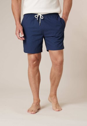 Homme portant un short bleu marine à cordon et une chemise blanche, debout pieds nus devant un fond uni.